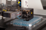 The photo shows a pipetting system above a multi-well cell culture plate placed on the microscope stage.<br /> <br />