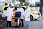 Three men in white coats stand behind a mobile stand equipped with several small devices.