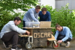 Vier Entwickler im Hintergrund basteln an Oscar im Vordergrund, dem Erntehelfer aus Holz mit Rädern.