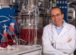 The photo shows a man with glasses and wearing a lab coat. The man has folded his arms and is laughing. There are three glass containes to his left, containing red liquids.<br />