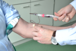Der Bildausschnitt zeigt den Arm eines Mannes (links), dem soeben Blut abgenommen wird. Über einen kleinen Schlauch fließt das Blut von der Kanüle in das Blutentnahmeröhrchen.