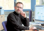 Das Foto zeigt einen dunkelhaarigen Mann mit Vollbart und Brille. Er trägt einen schwarzen Pullover und sitzt an einem Schreibtisch.