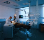 The photo shows a man and a woman in lab coats and face masks. Two doctors are bent over a man to examine his teeth. The patient is lying on a dentist's chair.