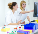 Zu sehen sind zwei Mitarbeiterinnen im Labor der Swiss Analysis AG, die an einem Labortisch sitzen, während eine der beiden Mitarbeiterinnen auf einen PC-Bildschirm zeigt.