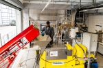 You can see a yellow machine with a funnel-shaped opening into which grassland cuttings are fed using a conveyor belt. A man stands behind the yellow machine and monitors the process. The biorefinery hall can be seen in the background.