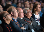Four men in suits listening to a speech.
