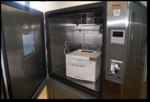 Zu sehen ist ein Schrank aus Metall, die Tür steht offen, in seinem Inneren steht ein weißer Laserdrucker.