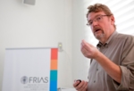 Zu sehen ist ein gestikulierender Mann mit Brille, im Hintergrund ein Poster mit dem Titel FRIAS