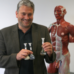 Bernhard Hirt, stehend vor einem anatomischen Modell eines Menschen, hält drei Glaskolben in den Händen.
