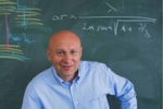 Zu sehen ist der Nobelpreisträger Stefan Hell, der verschmitzt grinsend vor einer Tafel steht.