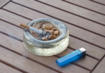 Eine brennende Zigarette liegt am Rand eines mit Kippen gefüllten Aschenbechers aus Glas.