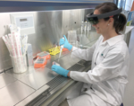 Eine Labormitarbeiterin in weißem Kittel und mit Datenbrille sitzt an
einer Sterilbank und pipettiert Flüssigkeit in ein Laborgefäß.