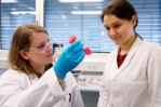 Zu sehen sind zwei junge Frauen in Laborkitteln, die sich eine rote Flüssigkeit im Plastikröhrchen anschauen.