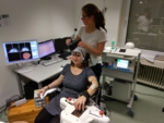 Female volunteer whose hand is connected to a robotic device that opens her hand while receiving magnetic stimuli via magnetic coils close to her head. The photo shows a member of Gharabaghi's team moving the magnetic coils over the volunteer's head.